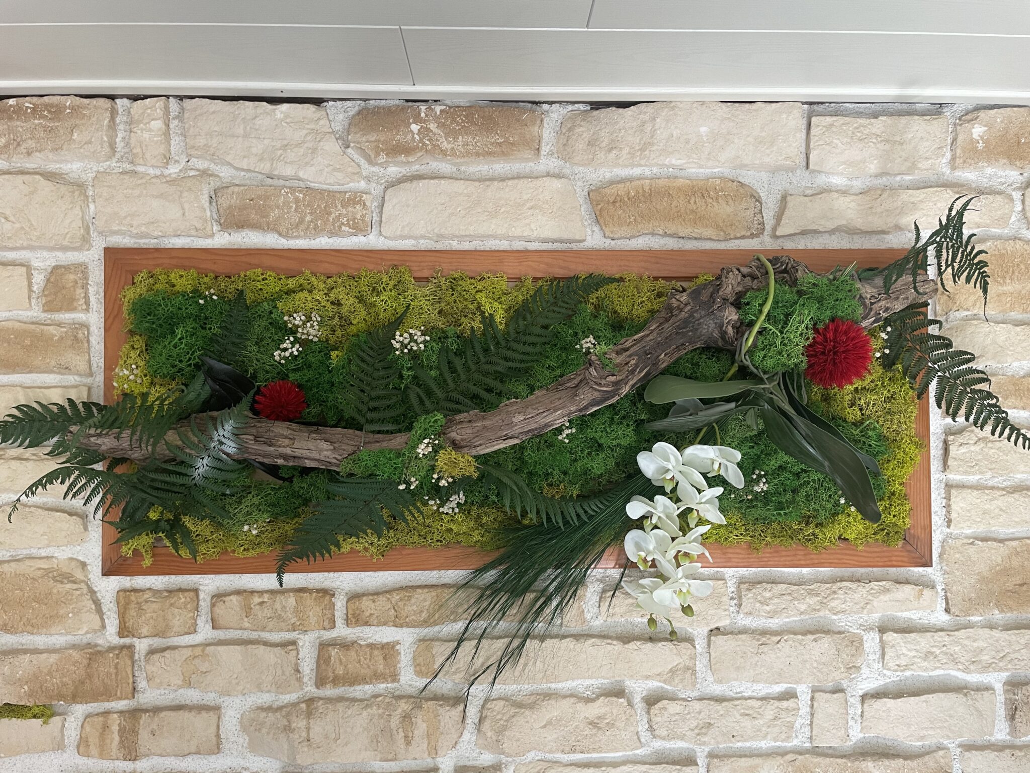 Composition florale murale avec mousse verte, branches, fougères et fleurs blanches et rouges sur un fond en pierre.